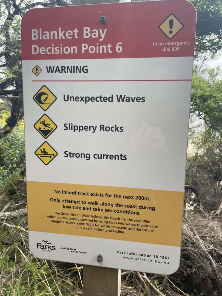 Warning sign at Blanket Bay advising against swimming due to unexpected rocks, slippery waves, and strong currents, and noting no Great Ocean Walk track for the next 200m.