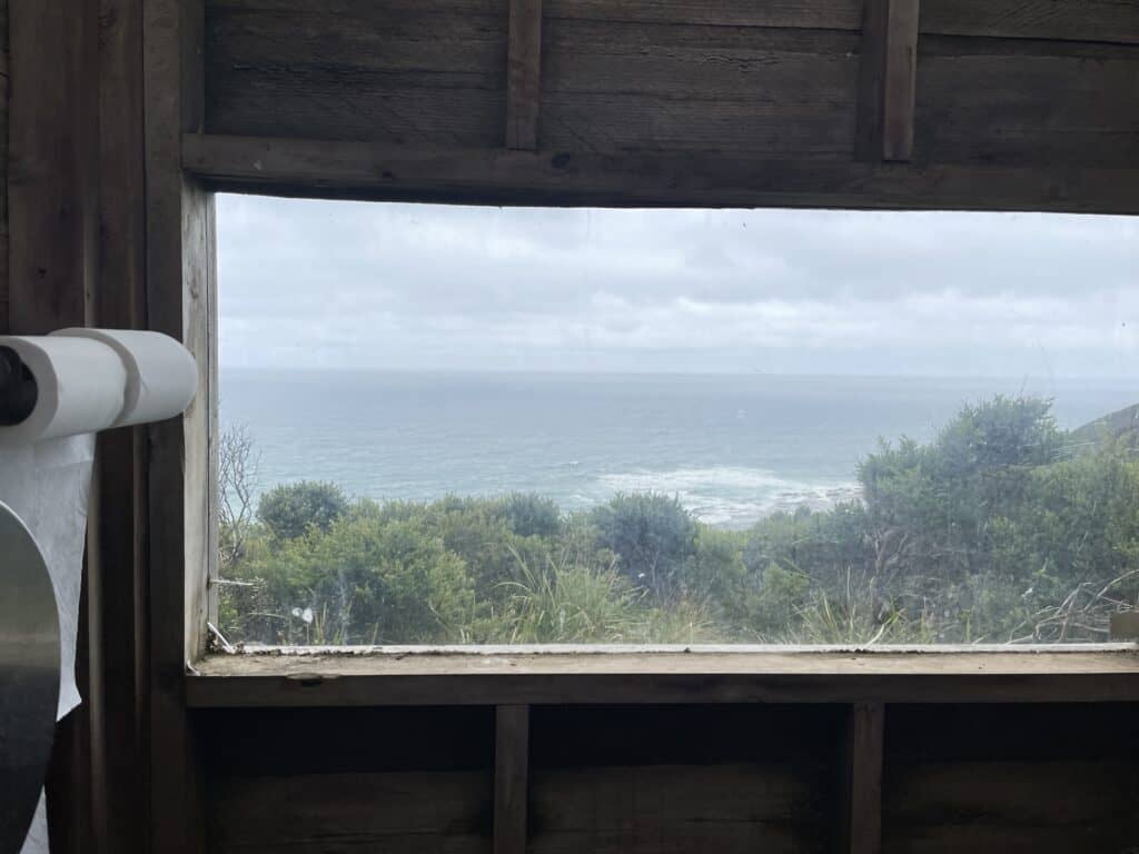 View of the ocean seen through a rustic wooden window with a visible roll of paper on the left, near one of the Great Ocean Walk campsites.