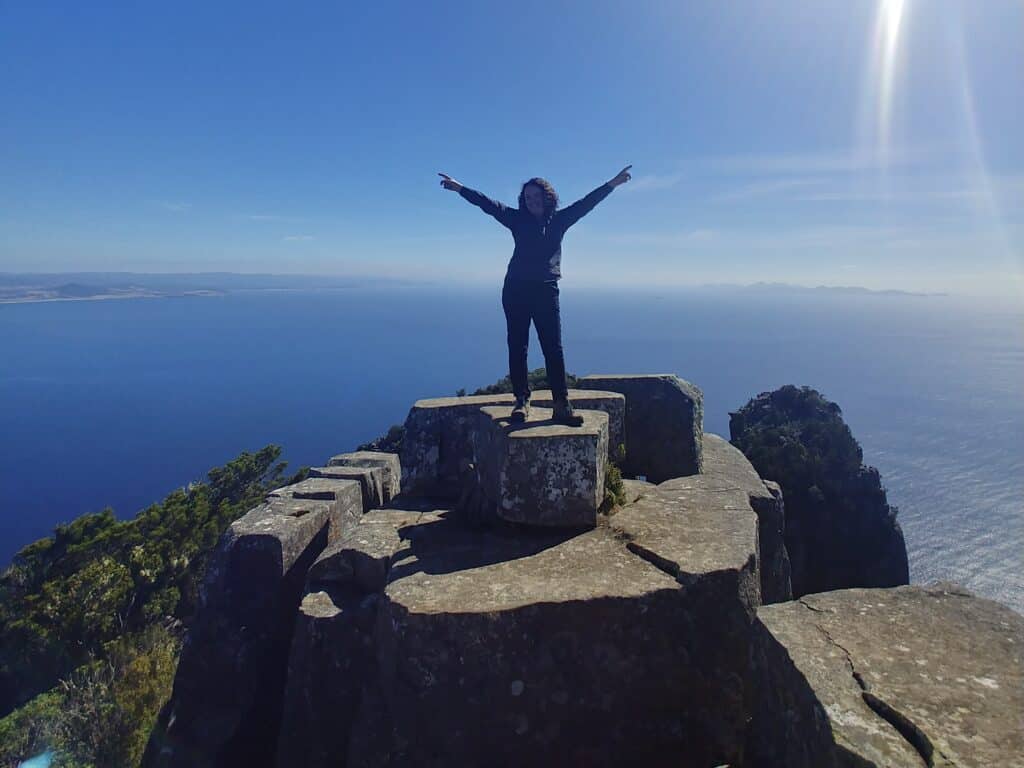 A person stands on a rocky outcrop with arms raised, overlooking an expansive ocean under a clear blue sky, capturing the essence of Maria Island hiking.