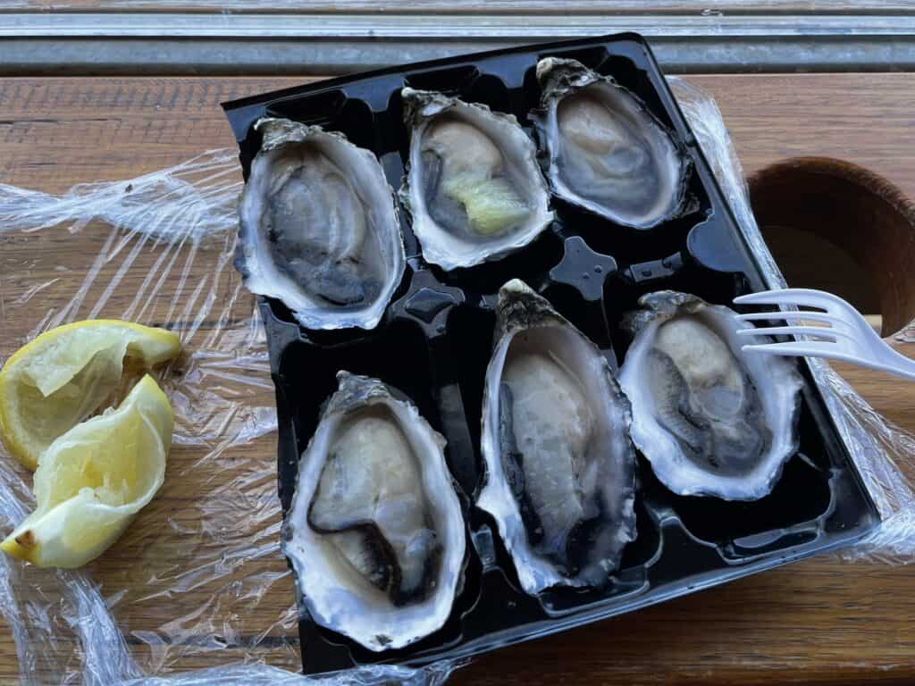 A plastic tray containing seven raw oysters on the half shell with a plastic fork and two squeezed lemon wedges on the side. The tray is placed on a wooden surface covered in plastic wrap, perfect for enjoying after a day of Maria Island hiking.