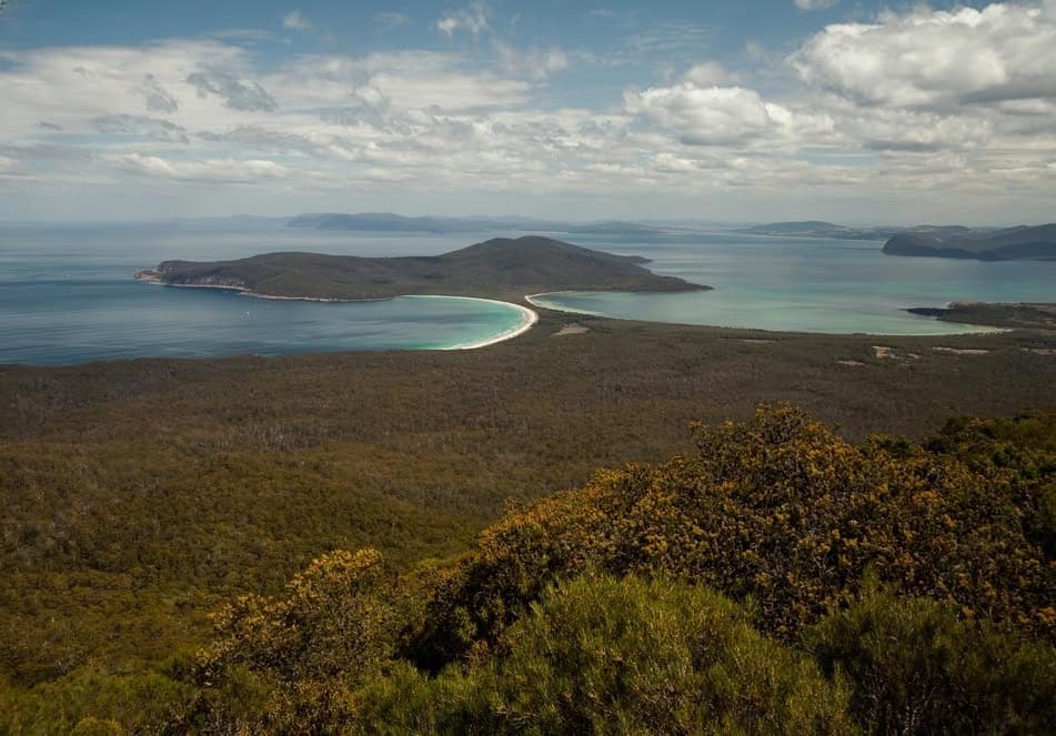 Aerial view of a forested peninsula on Maria Island, Tasmania, surrounded by the ocean with a curved sandy beach, under a partly cloudy sky. Perfect for hiking and exploring, with various Maria Island accommodation options nearby.