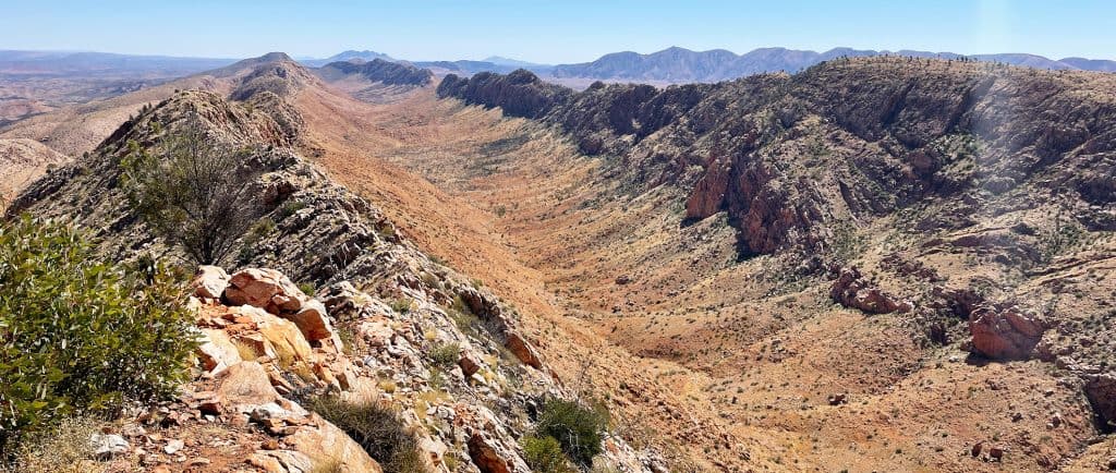 Panoramic view of a rugged mountain range along the Larapinta Trail, with sparse vegetation under a bright, clear sky.
