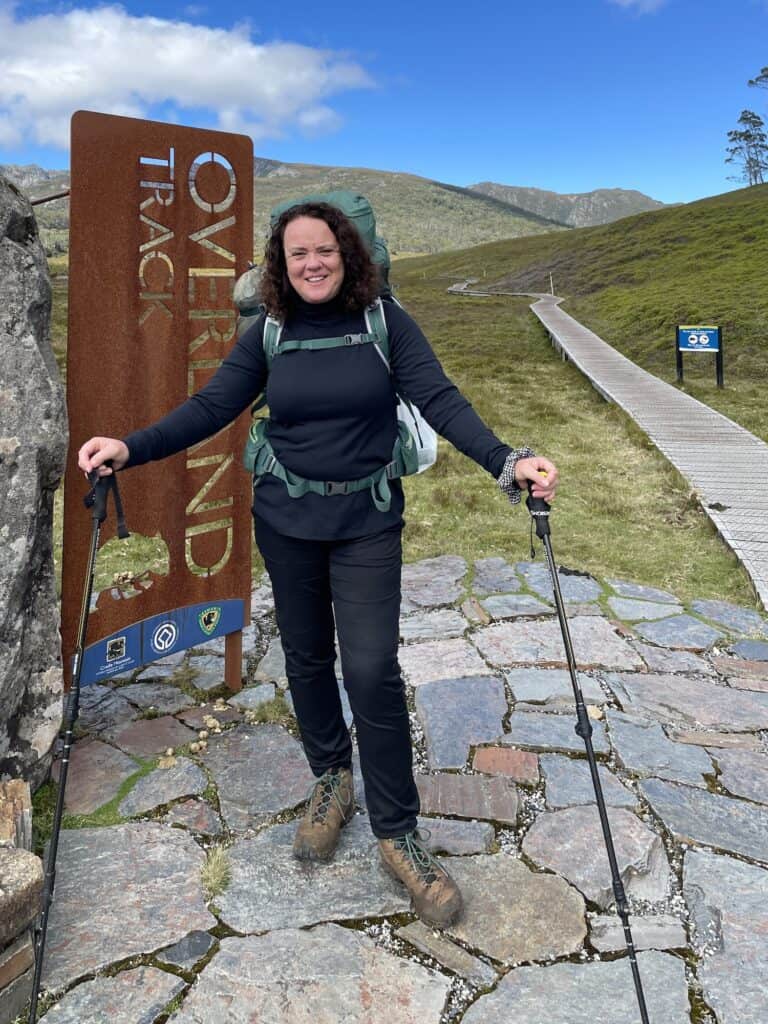 A woman in hiking gear stands at the start of the Overland Track solo, holding trekking poles. She is smiling and has a backpack on. The background shows a path leading into mountainous terrain.