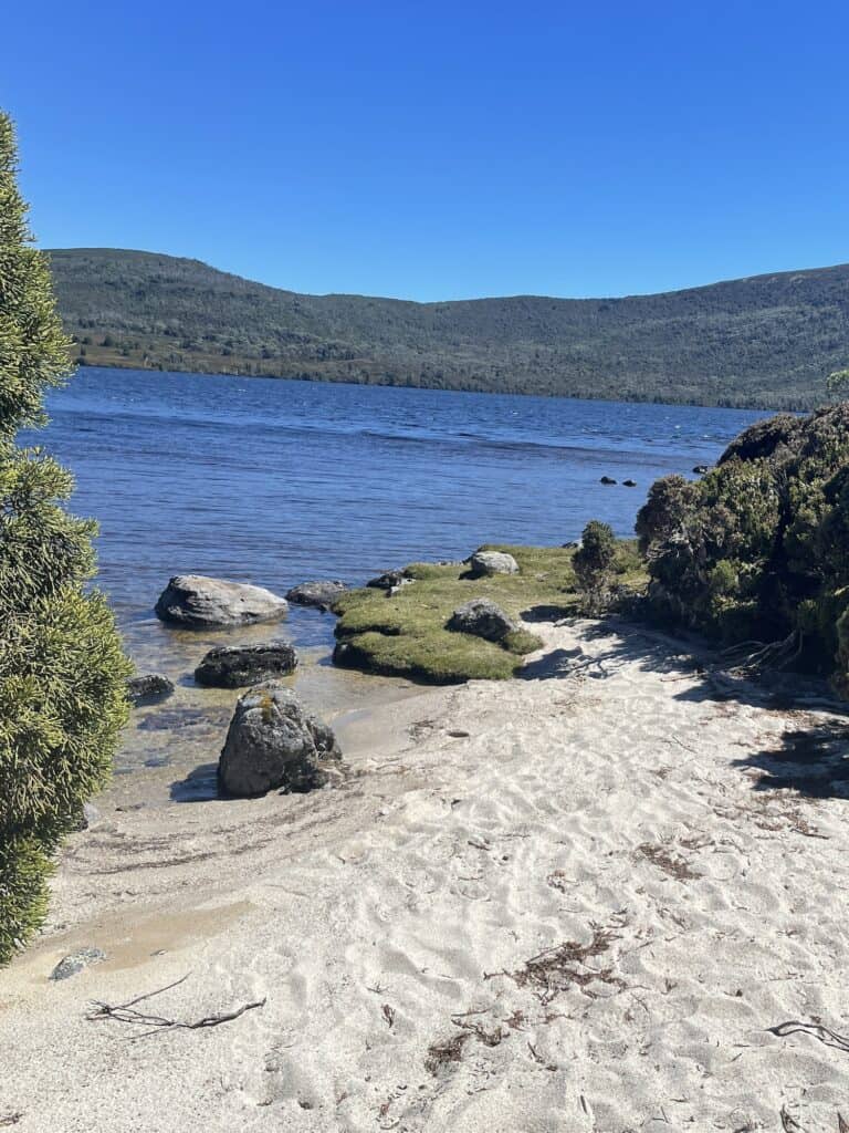 A sandy shoreline with scattered rocks and greenery leads to a clear blue lake set against a backdrop of green hills under a bright blue sky, perfect for those embarking on an overland track adventure.