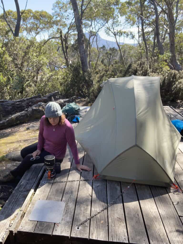 A person with a hat sits on a wooden platform beside a pitched tent in a forested area, preparing food on a camping stove, enjoying the serene beauty of their overland track accommodation.
