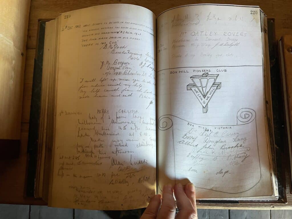 An open book showing handwritten text and drawings on both pages. The left page contains various signatures and notes, while the right page, perhaps from an overland track accommodation logbook, has a logo and a list of names and titles. A hand holds the book.