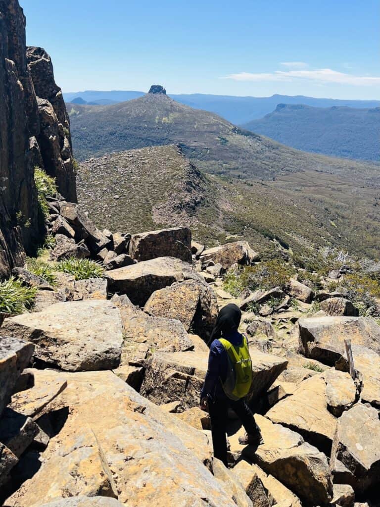 A person with a green backpack hikes over rocky terrain on the Never Never Trail, with a distant mountain peak under a clear blue sky.
