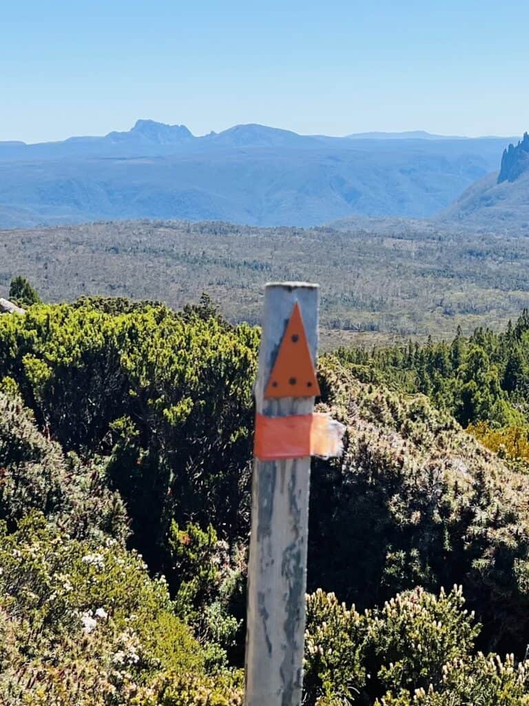 A wooden post with an orange triangular marker stands in the foreground of a scenic landscape featuring distant mountains and green vegetation under a clear blue sky, marking a trail often featured in the overland track packing list.