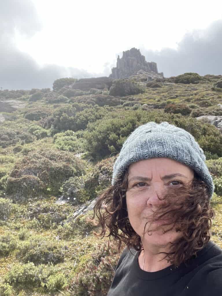 A person wearing a blue knit hat and black shirt stands outdoors with green vegetation and rocky hills in the background, possibly preparing for an adventure along the famous Overland Track.