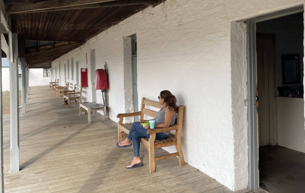 A person sits on a wooden bench on a covered wooden walkway beside a white brick building, holding a drink and looking into the distance, perhaps dreaming of their next Maria Island hiking adventure in Tasmania.