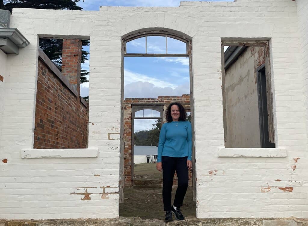 A person in a blue sweater and black pants stands in the doorway of a partially ruined brick and white plaster building, smiling at the camera, perhaps pondering their next Maria Island hiking adventure.