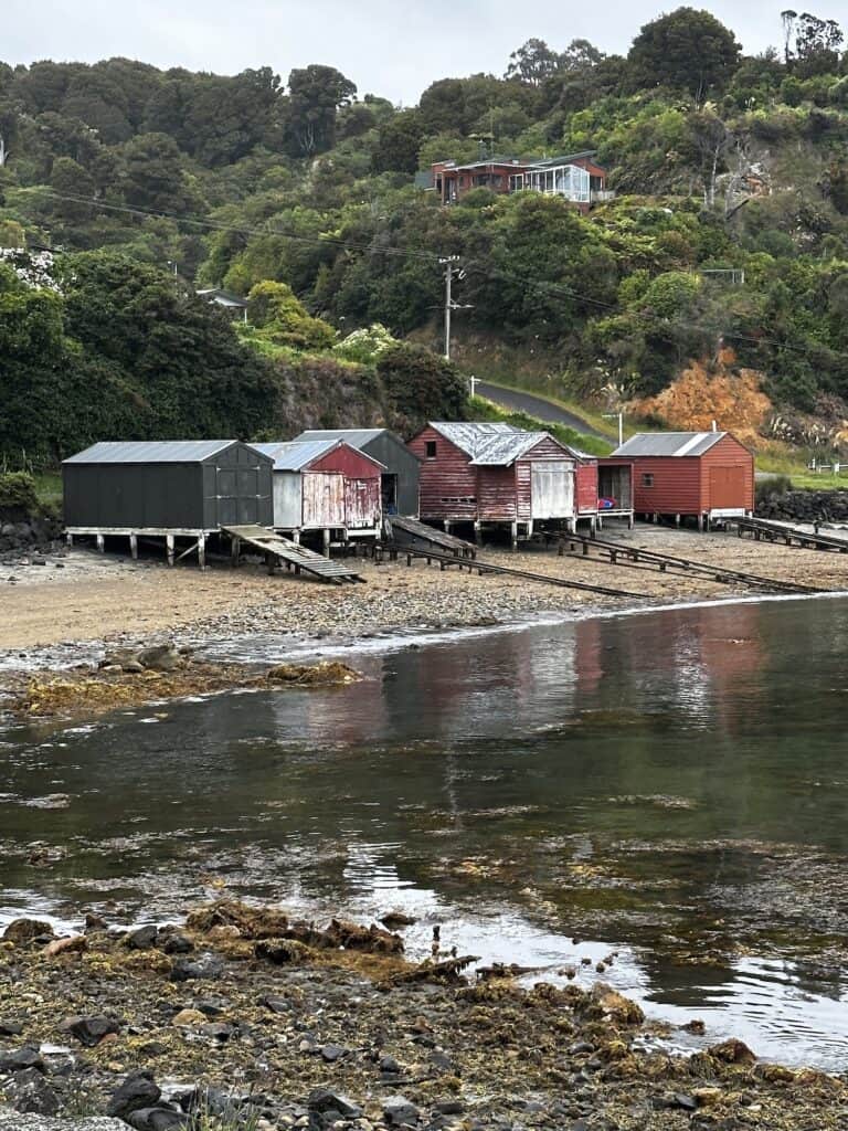 A row of small, colorful boathouses stands on a rocky shore with lush green hills and scattered houses in the background. Calm waters reflect parts of the scene.