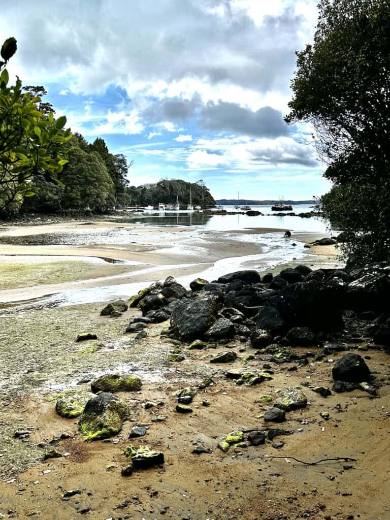 A beach with rocks and low tide, bordered by trees. Small boats are anchored in the distance on the water. Cloudy sky above.