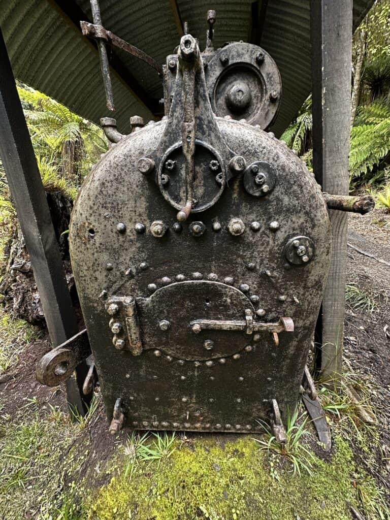 A rusted, black metal boiler with rivets and bolts stands under a corrugated metal canopy in an outdoor setting surrounded by greenery.
