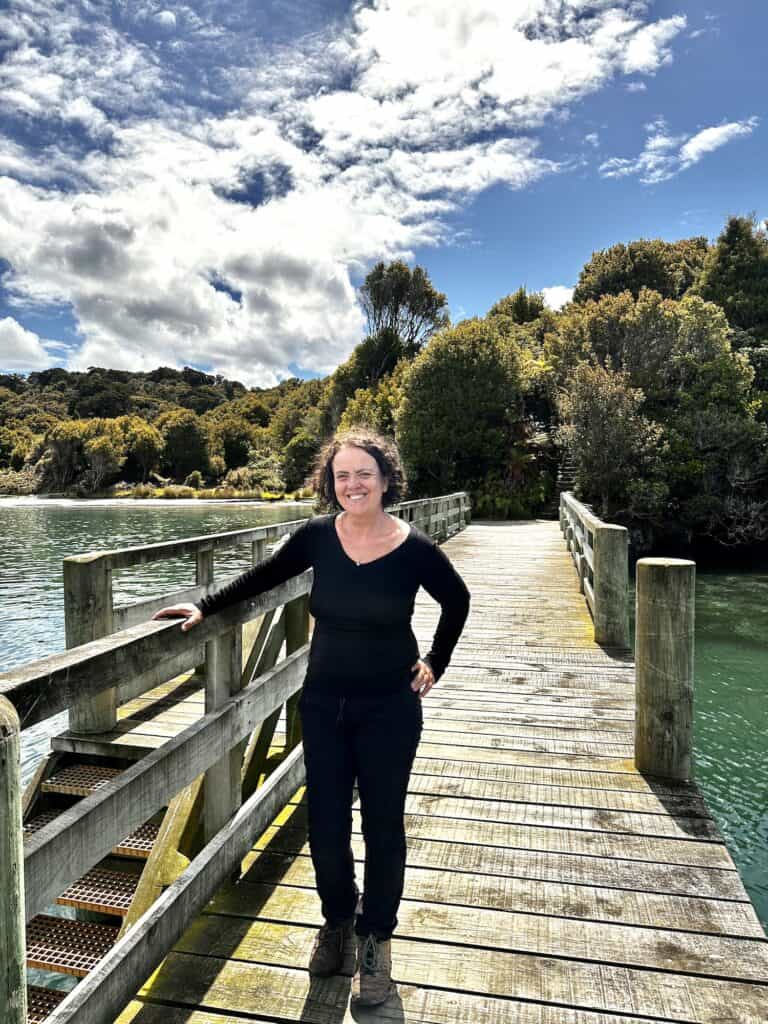 A woman in black clothing stands on a wooden bridge over a body of water, surrounded by trees under a partly cloudy sky.