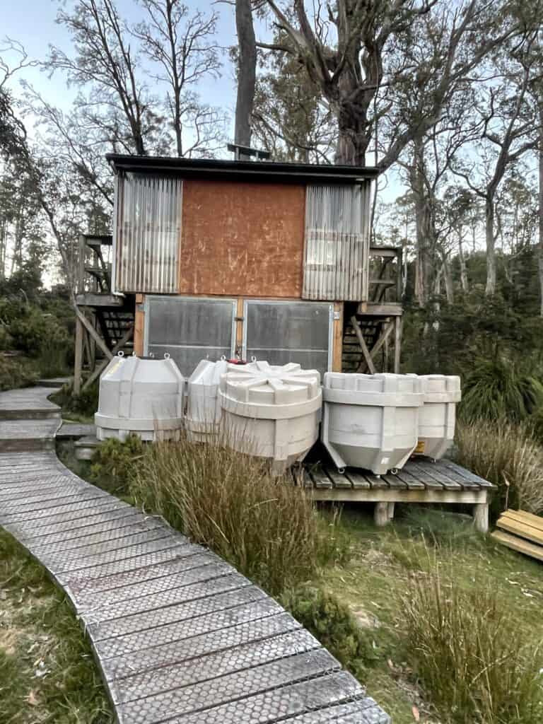 A wooden building on stilts surrounded by trees, with large, secured translucent storage containers in front, and a wooden boardwalk leading up to it—perfect for those seeking unique overland track accommodation.