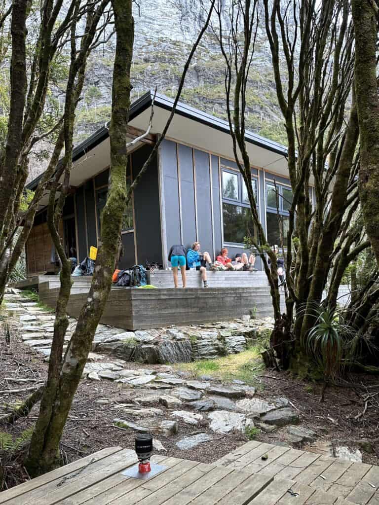 A group of people gather on the wooden porch of a modern cabin surrounded by tall, thin trees on a rocky mountainside, discussing their Frenchmans Cap packing list for the upcoming hike.