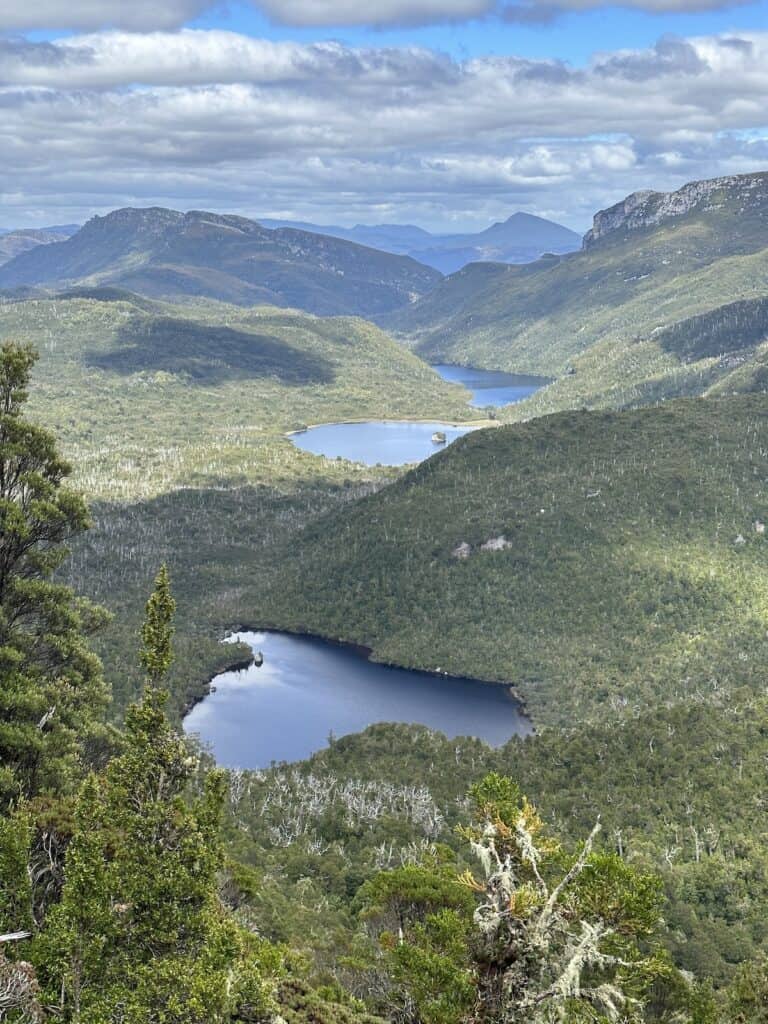 Aerial view of a lush green valley with multiple serene blue lakes surrounded by mountains under a partly cloudy sky, perfect for adventurers ready to tackle the Frenchmans Cap.