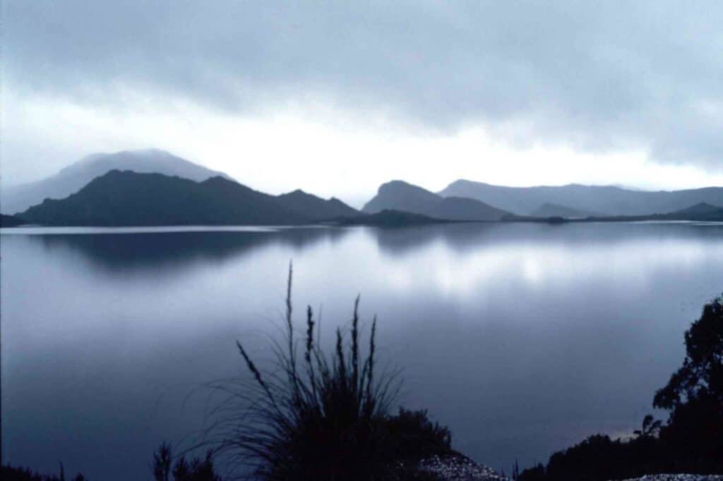 Serene lake reflecting dark, cloudy sky with distant mountains and foreground vegetation, reminiscent of the tranquil views en route from Hobart to Derwent Bridge.