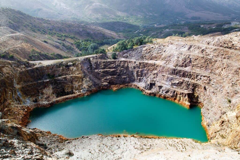 A large turquoise-colored mining pit filled with water, surrounded by rocky terrain and hills in the background, reminiscent of the scenic landscapes you'd encounter when traveling from Queenstown to Derwent Bridge.