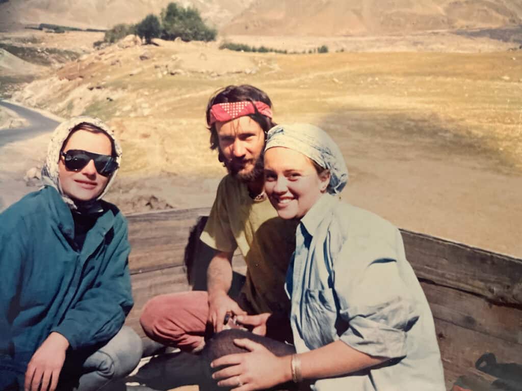 Three people sitting outdoors on a wooden structure in a mountainous landscape, with two wearing headscarves and one wearing sunglasses and a bandana.