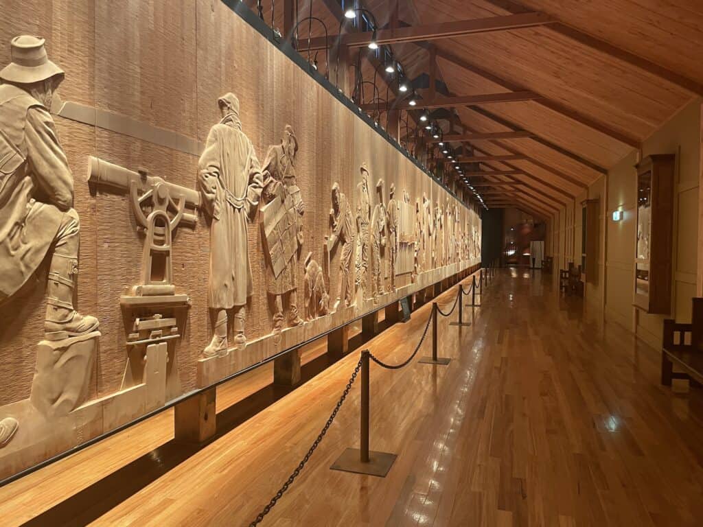 A long wooden relief sculpture depicting various historical figures engaged in different activities is mounted on a wooden wall in a room with polished hardwood floors and track lighting, reminiscent of the rich history you'll find when traveling from Hobart to Derwent Bridge.