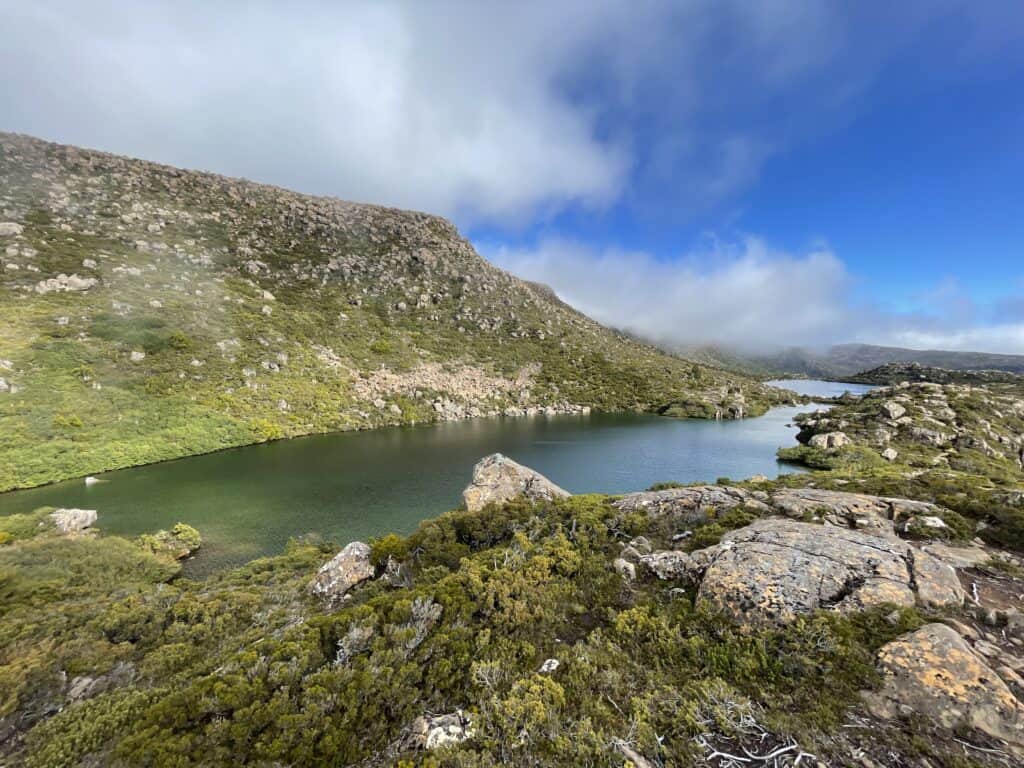 A scenic view of a mountain tarn in Mt Field National Park surrounded by rocky terrain and green shrubbery under a partly cloudy sky.
