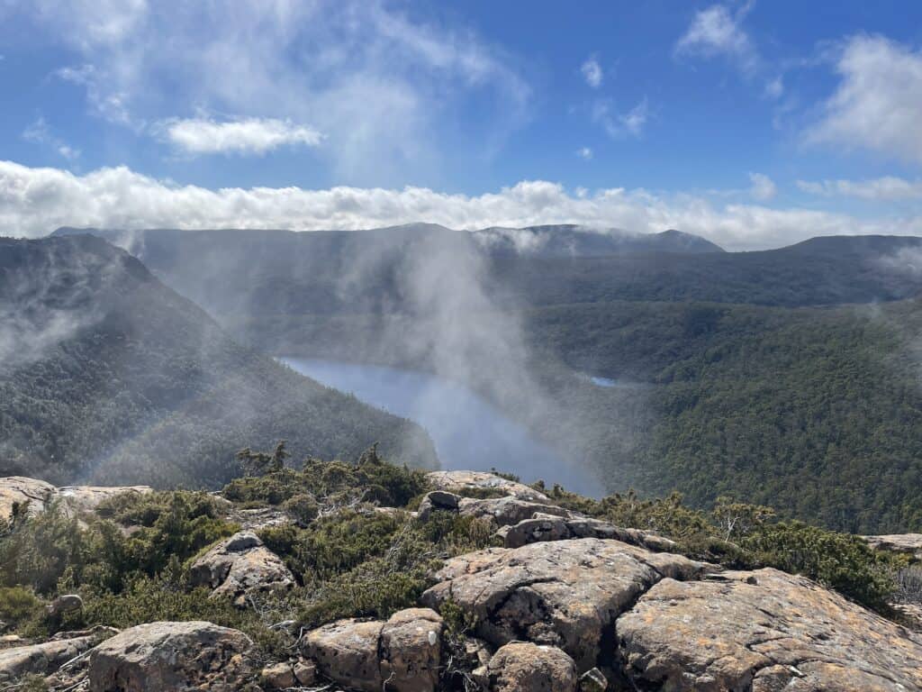 A landscape view featuring a rocky foreground, a forested valley, and a serene lake, with mist and clouds partially obscuring the distant hills under a blue sky on the Tarn Shelf Circuit