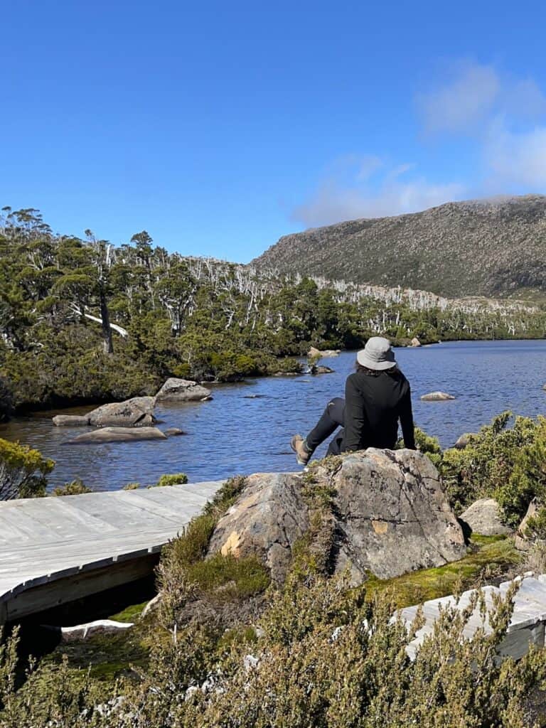 A person in a black jacket and gray hat sits on a rock by a lake, with a wooden footbridge and lush greenery in the foreground and the stunning Tarn Shelf under a clear blue sky in the background.