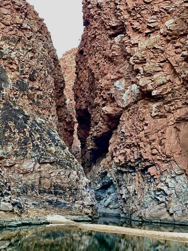 Narrow gorge with steep, rugged red cliffs along the Larapinta Trail and a calm waterway at the base, reflecting the rocky walls.