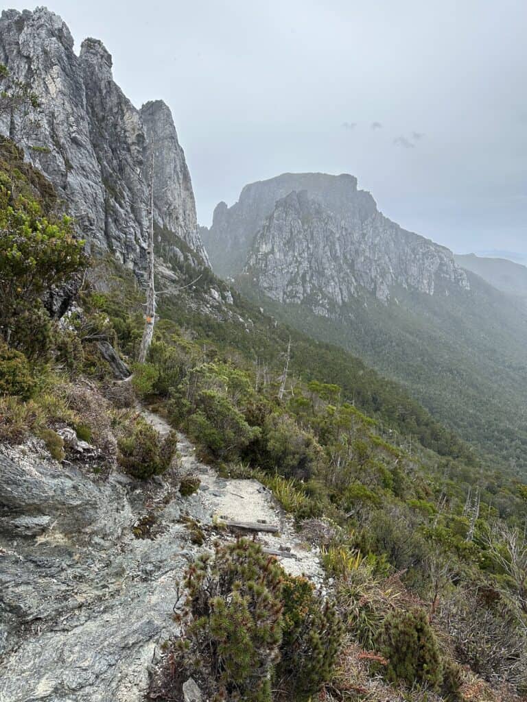 A rocky trail winds through a mountainous landscape with steep cliffs and dense greenery, similar to the terrain you'll find on the journey from Strahan to Derwent Bridge. Mist or fog adds a touch of fogginess to the distant mountain peaks, reminiscent of views from Iron Blow Lookout.