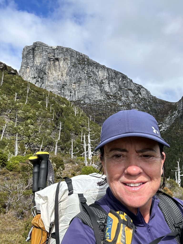 A person in a blue cap and outdoor gear stands in front of a rocky cliff and forested hillside, smiling at the camera, likely near the scenic Iron Blow Lookout on their journey from Queenstown to Derwent Bridge.