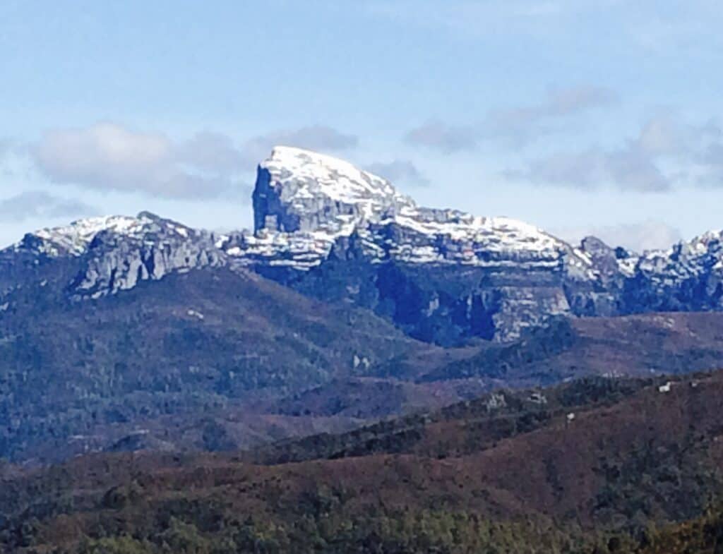 Mountain range with snow-capped peaks under a partly cloudy sky, viewed from a distance. Trees and hills in the foreground transition into rocky, elevated terrain near Queenstown to Derwent Bridge.