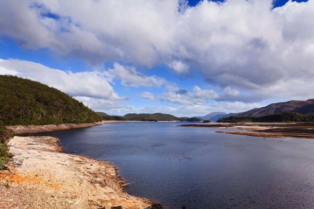 A serene lake surrounded by forested hills under a partly cloudy sky, reminiscent of the views you might see on the drive from Derwent Bridge to Strahan. The lake's shoreline shows a contrast of rocky and vegetated areas.
