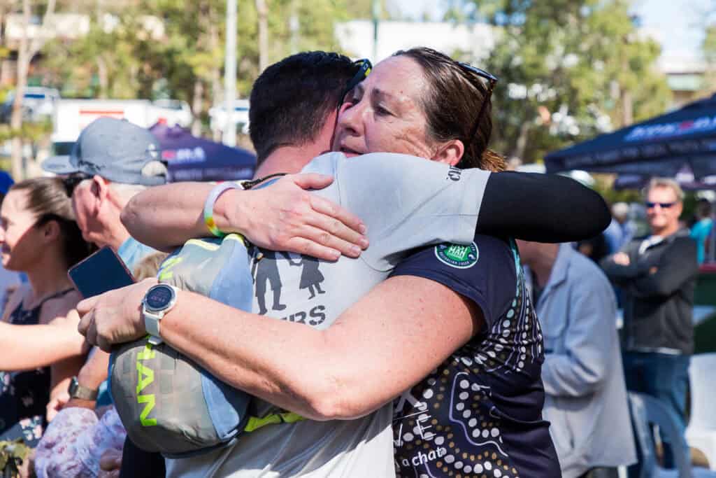Two people embrace tightly in an outdoor setting, surrounded by others. Both wear event T-shirts, and one person's back is visible, displaying graphics and text. They are part of the Kokoda Challenge support crew, providing encouragement and strength to all participants in this monumental event.