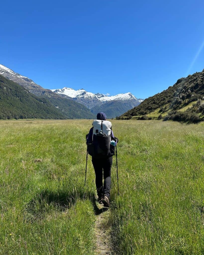 A person with a large backpack and hiking poles walks through a grassy field towards distant snow-capped mountains under a clear blue sky.
