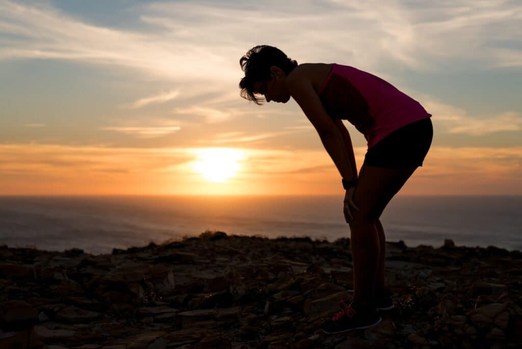A person in athletic wear bends forward, resting hands on knees, against a sunset backdrop on a rocky terrain near the ocean. This scene is perfect for sharing tips for hiking in hot weather to ensure you stay safe and enjoy your summer adventures.