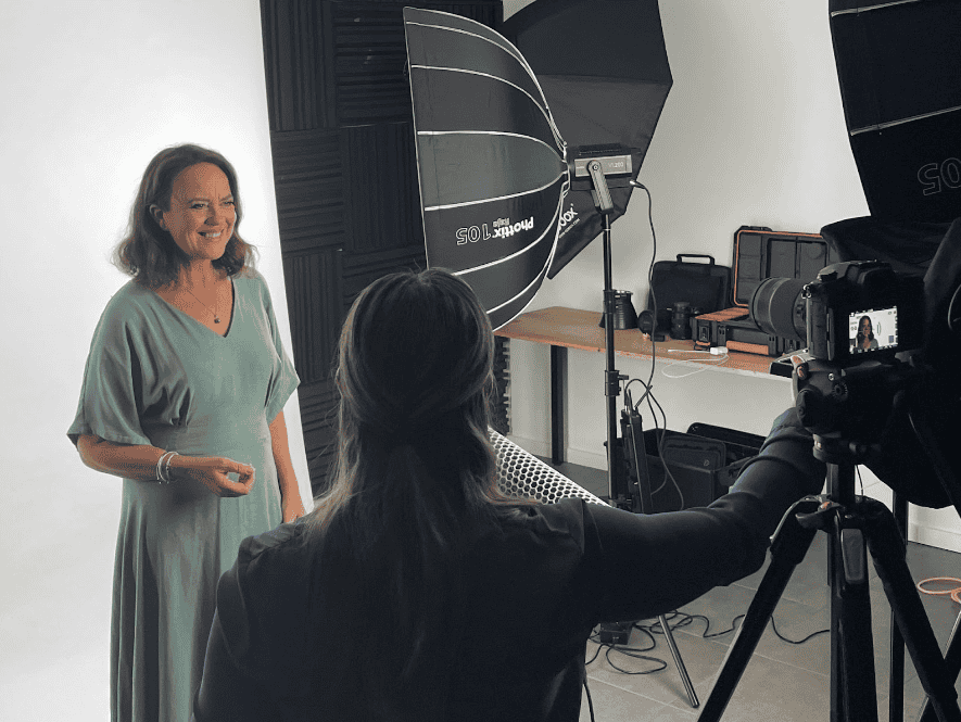 A woman in a green dress is standing in a photography studio, posing for a photoshoot with a photographer capturing her image. Studio lighting and equipment are set up around them.