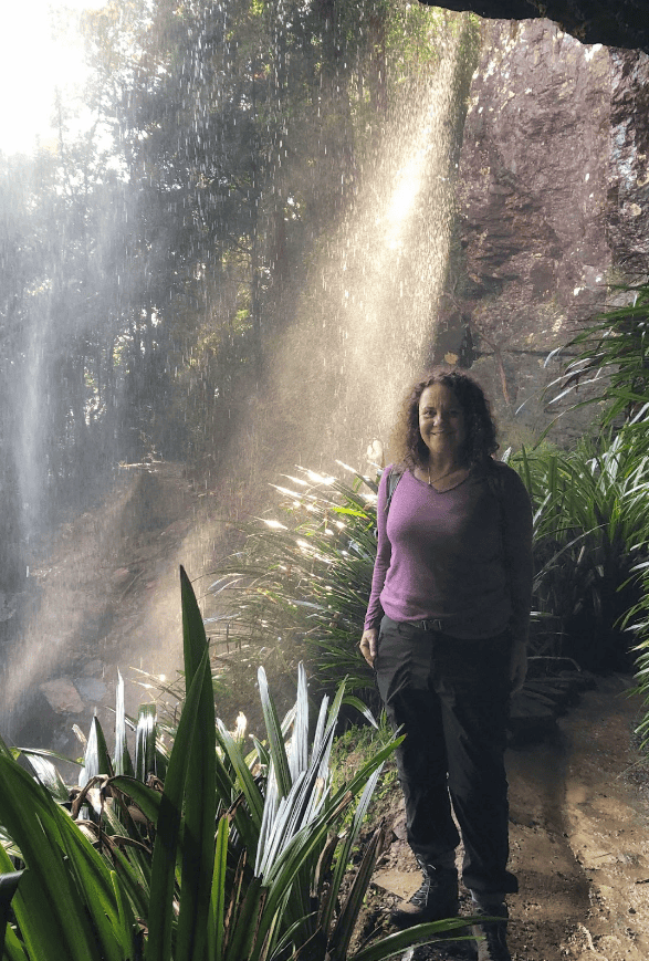 A woman stands on a narrow path under a waterfall, surrounded by lush greenery, with sunlight filtering through the water.