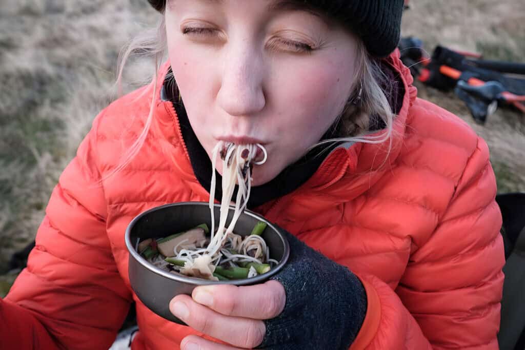 Person in a red jacket and black beanie eating noodles from a bowl outdoors, providing much-needed energy to the Kokoda Challenge support crew.