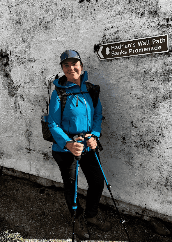 Person in a blue jacket and cap stands near a sign reading "Hadrian's Wall Path Banks Promenade," holding trekking poles.