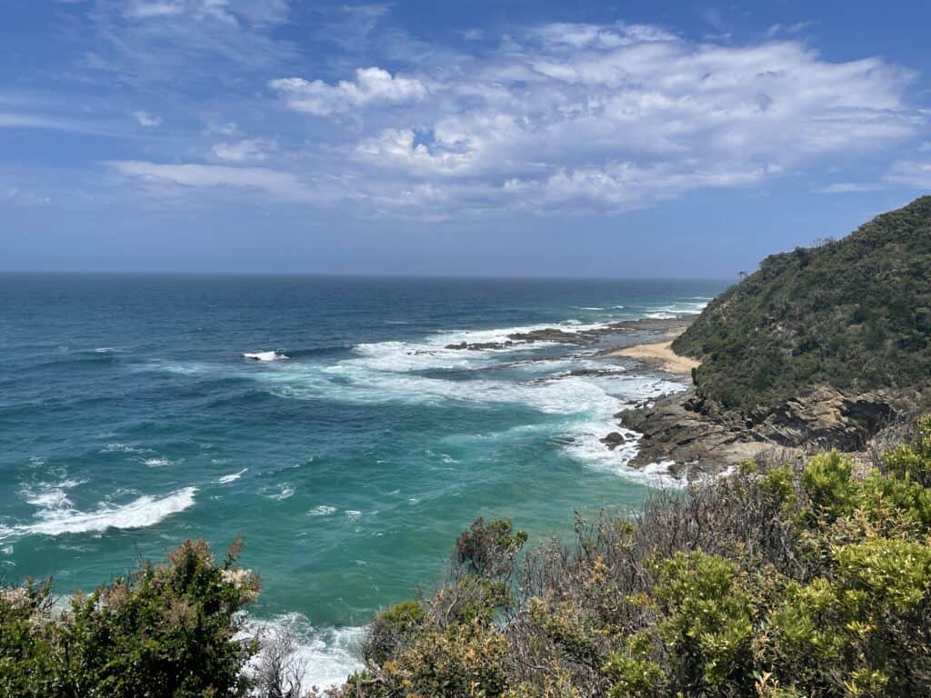 Coastal view showing turquoise waves crashing against rocky shores under a blue sky, with lush green foliage in the foreground, perfectly capturing the essence of hiking the Great Ocean Road.