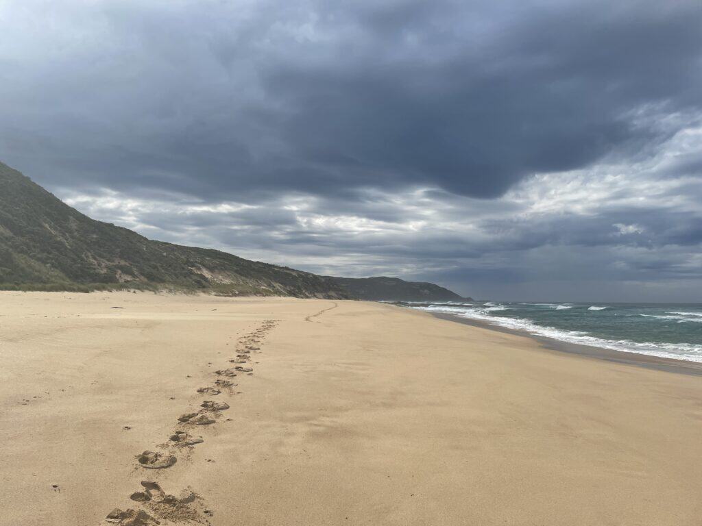Footprints on a sandy beach leading toward distant hills under a cloudy sky along the Great Ocean Walk.