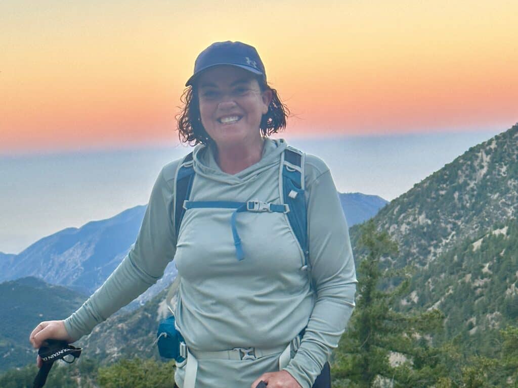 A woman in hiking gear smiles at the camera with a mountainous landscape and a sunset in the background.