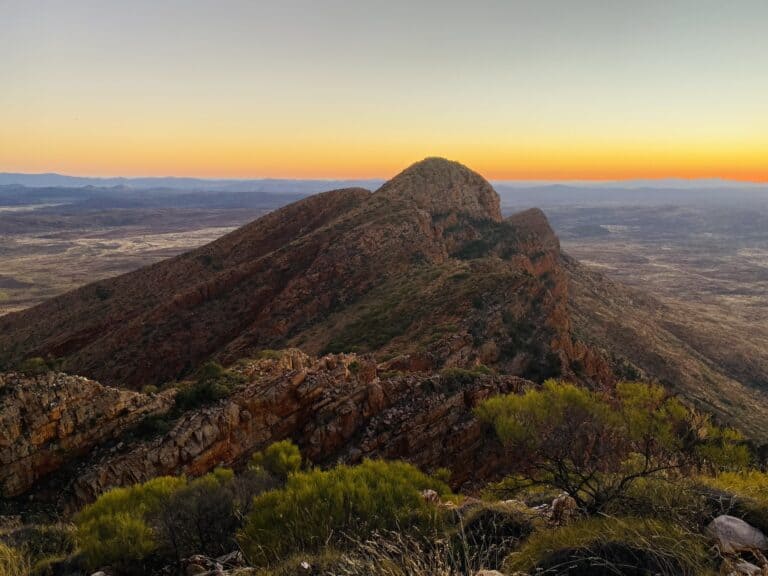 Sunset casting warm hues over a rugged mountain range with a distinct, dome-shaped peak and expansive views of the horizon near Redbank Gorge.
