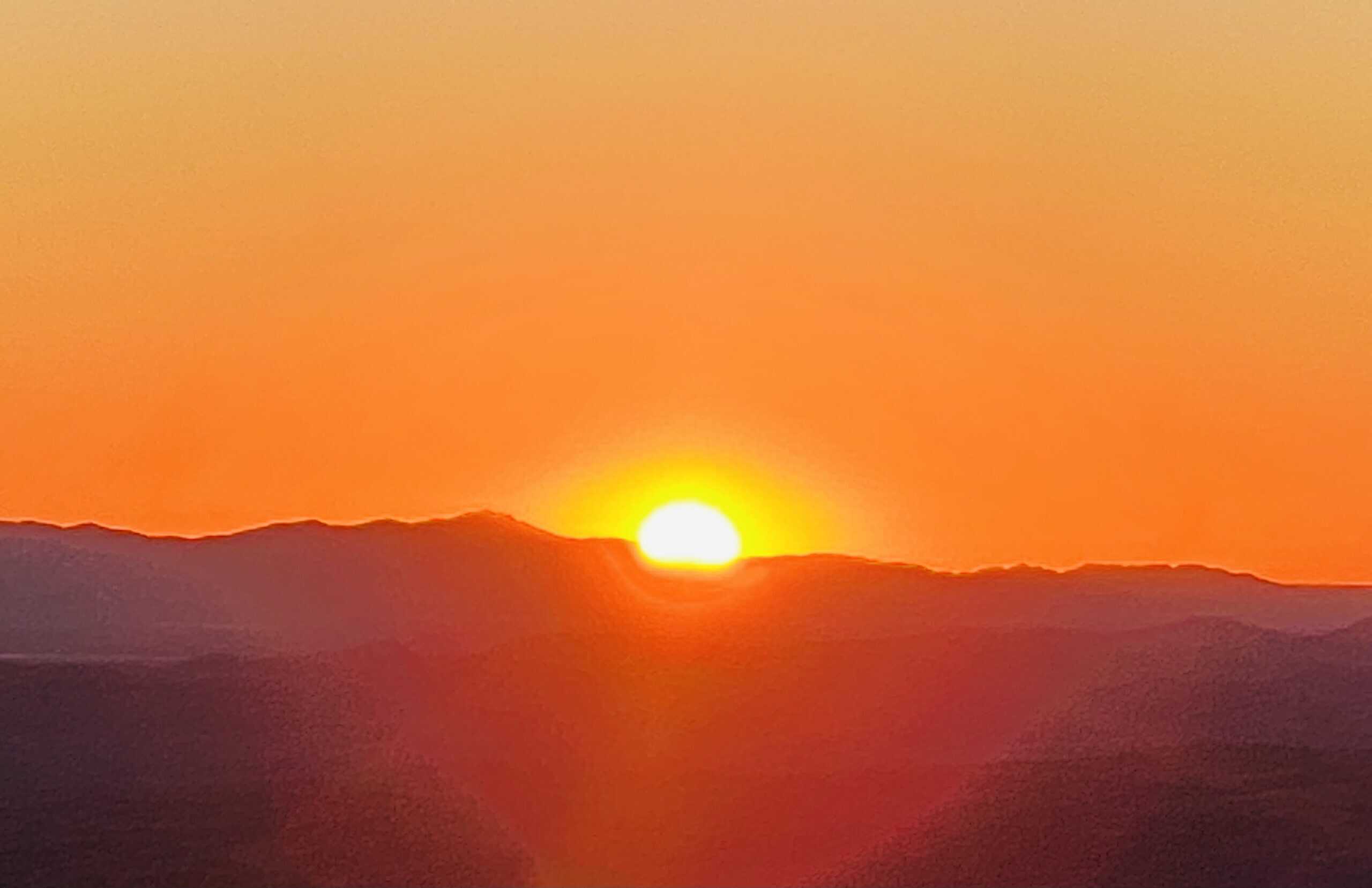 A sun is setting behind a range of mountains, casting an orange and yellow glow in the sky and creating dark silhouettes of the peaks. As you journey along the Larapinta Trail for 14 days, evenings like this offer a serene transition from day to dusk, often enjoyed after savoring Larapinta Trail food.