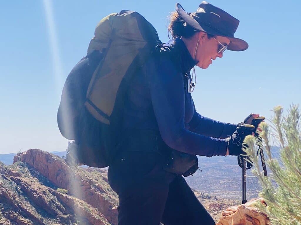 A female hiker with a large backpack and trekking poles ascends a rocky trail, wearing sunglasses and a hat, with a clear sky above.
