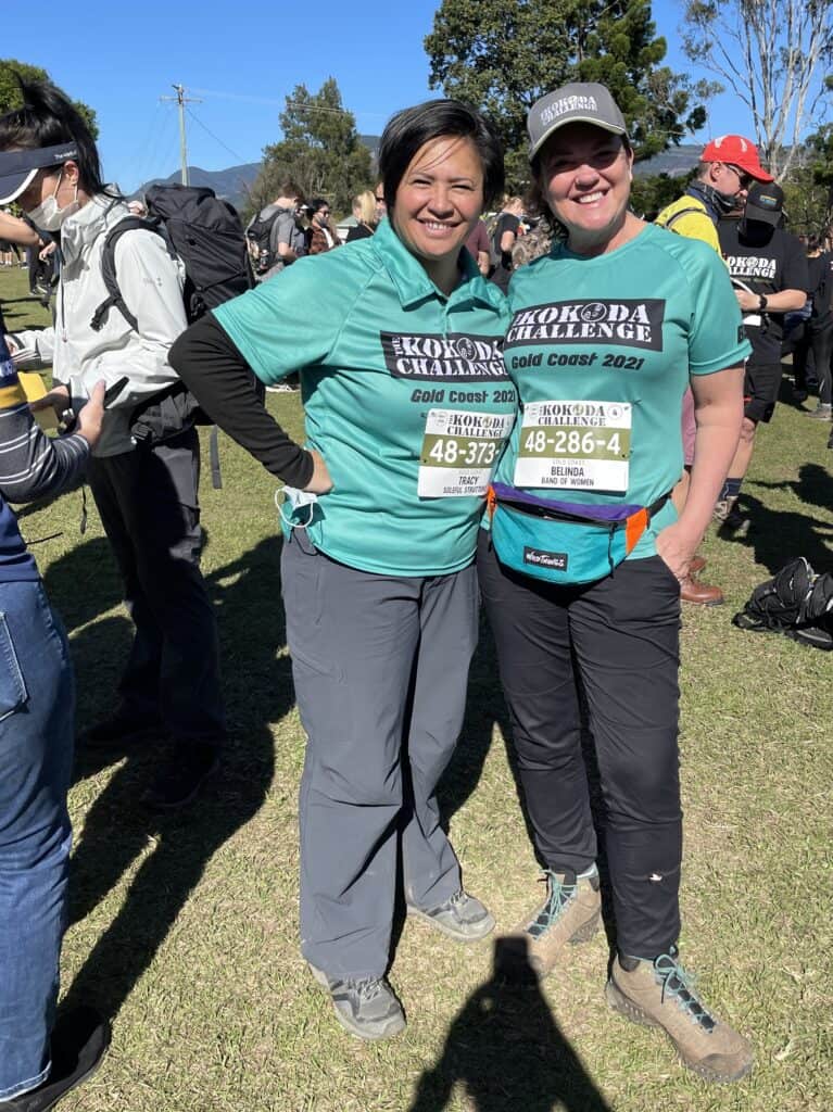 Two women wearing teal Kokoda Challenge 2021 shirts and race bibs pose for a photo outdoors, showcasing their kokoda challenge preparation. Other participants can be seen in the background.
