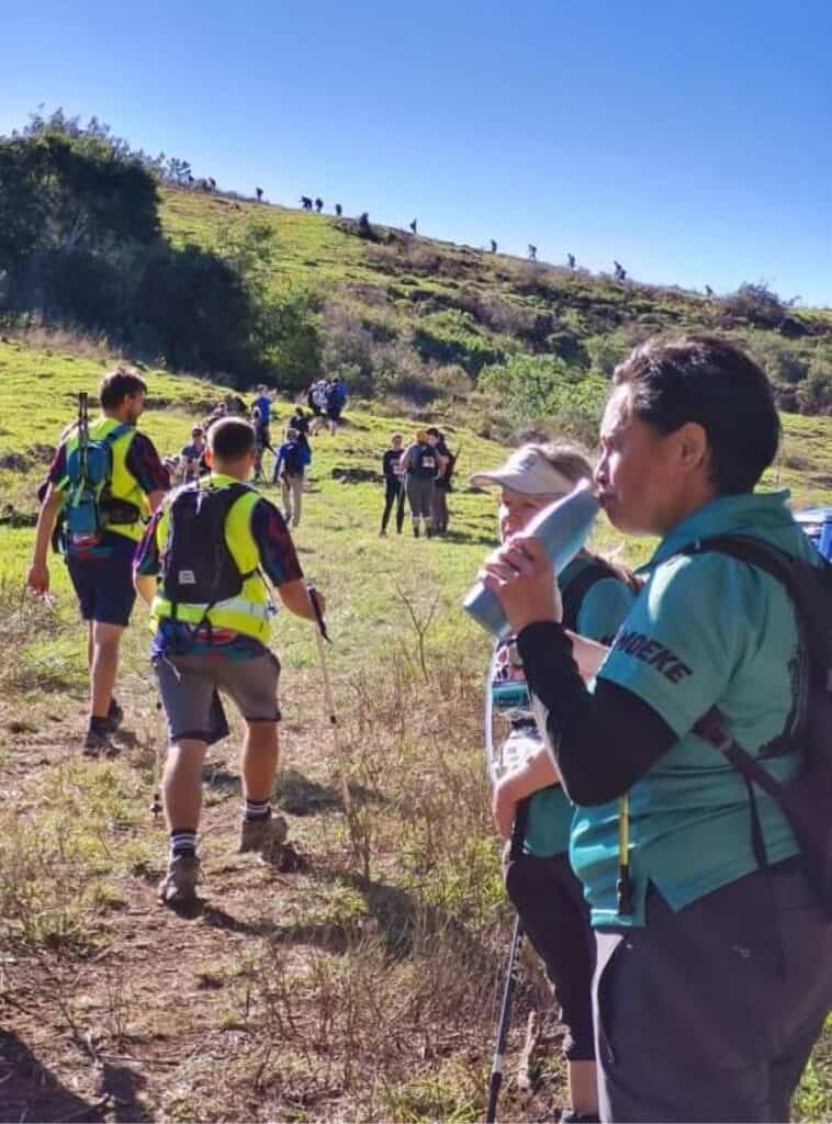 Hikers traverse a grassy hillside under a clear blue sky, with some resting and others continuing up the path, preparing for the Kokoda Challenge. A person in the foreground drinks from a bottle.
