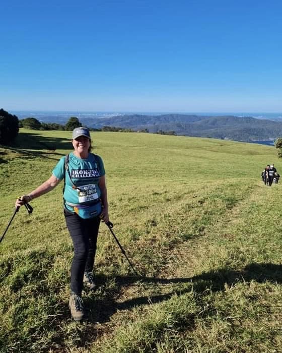 A person with hiking poles stands on a grassy trail with a mountainous landscape and clear blue sky in the background, likely preparing for the Kokoda Challenge. Other hikers are visible in the distance, perhaps discussing their Kokoda Challenge gear.